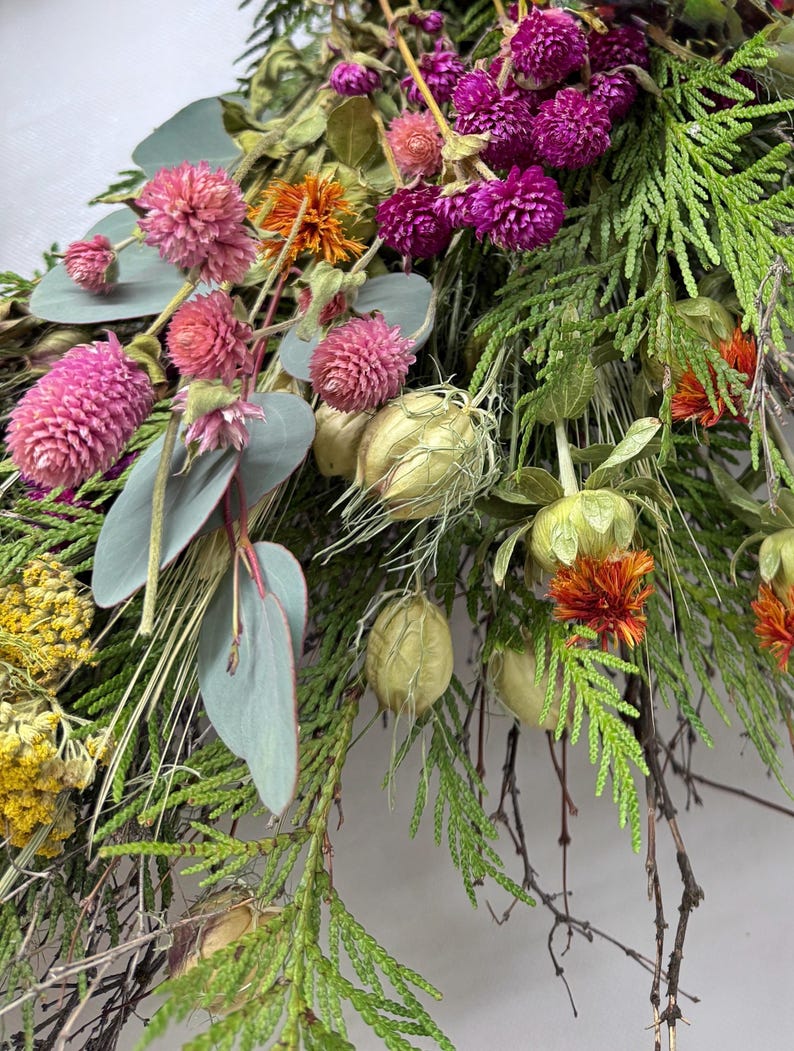 K&ouml;nnte beinhalten: Nahaufnahme eines getrockneten Blumenarrangements mit rosa Kugelamarant, lila Gomphrena, orangefarbenen Bl&uuml;ten und gr&uuml;nem Laub. Das Arrangement umfasst Eukalyptusbl&auml;tter und Samenkapseln und erzeugt eine nat&uuml;rliche, strukturierte &Auml;sthetik.