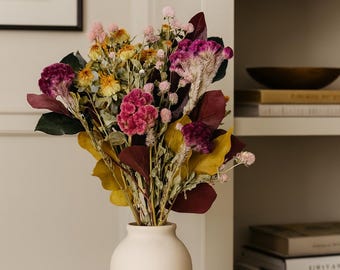 May include: A white ceramic vase holds a vibrant bouquet of dried flowers. The arrangement includes yellow, pink, and burgundy blooms with green and burgundy leaves. The vase sits on a white surface, with a framed picture and a bookshelf in the background.