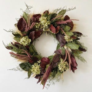 May include: A circular wreath composed of dried botanical elements, featuring deep burgundy leaves, pale green hydrangea clusters, feathery pink amaranth plumes, and stalks of golden wheat arranged in a dense, textured, and layered circular pattern.