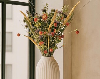May include: A white, ribbed ceramic vase filled with a bouquet of dried flowers. The arrangement includes orange, purple, and green elements, with tall, wheat-like stalks. The vase sits on a white table near a window.