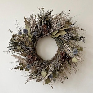 May include: A circular wreath constructed from dried botanical elements including brown pinecones, muted green eucalyptus leaves, and textured blue globe thistle heads. The arrangement features a variety of dried grasses, twigs, and foliage.