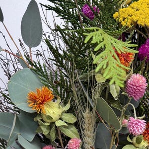 K&ouml;nnte beinhalten: Nahaufnahme eines Trockenblumenarrangements mit verschiedenen Texturen und Farben. Enth&auml;lt gr&uuml;nes Laub, orange, gelbe und rosa Bl&uuml;ten sowie getrocknete Weizen&auml;hren. Die Anordnung befindet sich vor einem wei&szlig;en Hintergrund.