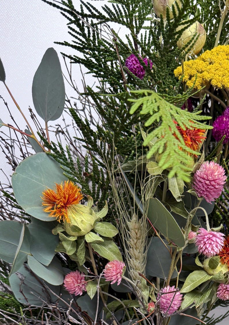 K&ouml;nnte beinhalten: Nahaufnahme eines Trockenblumenarrangements mit verschiedenen Texturen und Farben. Enth&auml;lt gr&uuml;nes Laub, orange, gelbe und rosa Bl&uuml;ten sowie getrocknete Weizen&auml;hren. Die Anordnung befindet sich vor einem wei&szlig;en Hintergrund.