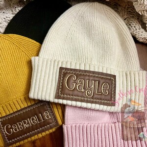 May include: Three knitted beanies with personalized leather patches. The beanies are in mustard yellow, cream, and pink. The patches are brown with white stitching and the names "Gabriella", "Gayle", and "Southern Craft" are embroidered on them.
