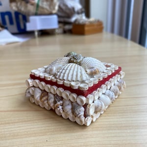 May include: A square jewellery box decorated with various seashells. The box has a red velvet lining and is adorned with small, white seashells around the edges. The top is covered with larger, white seashells.