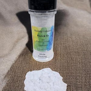 May include: A small white jar of flocking material labelled "Flocking Unlimited Flock & Go (1oz) Flocking Fibers and Applicator". The jar is sitting on a brown fabric surface with a small pile of white flocking material next to it.