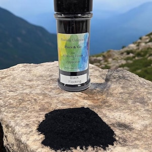 Puede incluir: Un pequeño frasco de material de flocado negro con una etiqueta que dice "Flocking Unlimited Flock & Go (1oz) Flocking Fibers and Applicator". El frasco está sentado en una roca con un montón de material de flocado negro en frente.