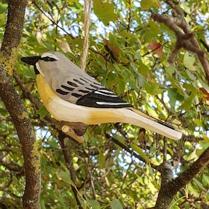 Puede incluir: Adorno de pájaro de madera tallada a mano, pintado en tonos grises, amarillos, negros y blancos, colgado de una cuerda de hilo natural. El pájaro está posado en una pequeña rama de madera, con un diseño detallado de alas. El fondo muestra ramas de árboles y hojas verdes.