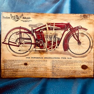 May include: Vintage wood sign advertising a 1918 Indian Powerplus motorcycle, model N-18. The sign features a detailed illustration of the motorcycle in red and black, with the words "Indian Motorcycles" and "1918 Powerplus Specifications - Type N-18" printed on the sign.