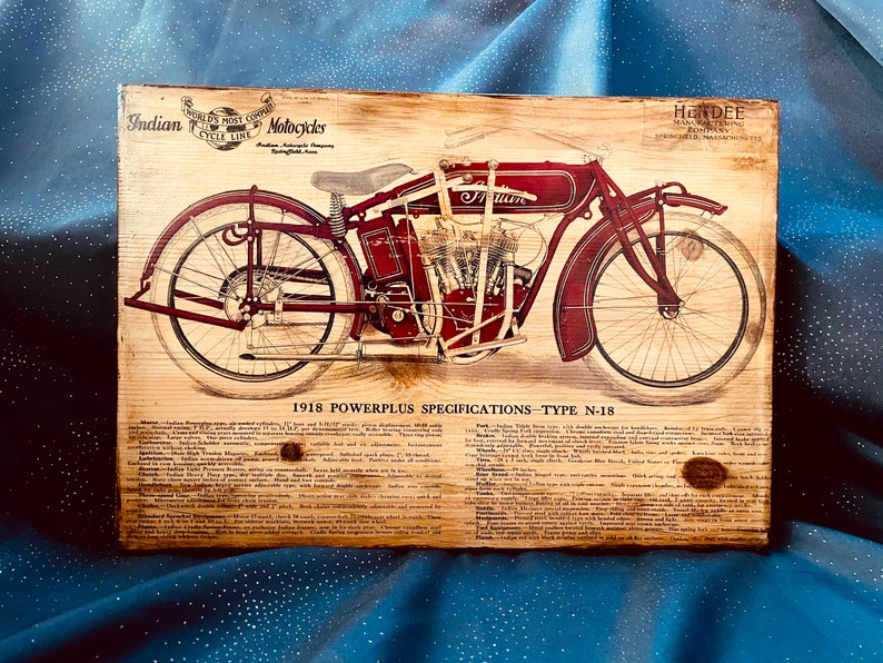May include: Vintage wood sign advertising a 1918 Indian Powerplus motorcycle, model N-18. The sign features a detailed illustration of the motorcycle in red and black, with the words "Indian Motorcycles" and "1918 Powerplus Specifications - Type N-18" printed on the sign.