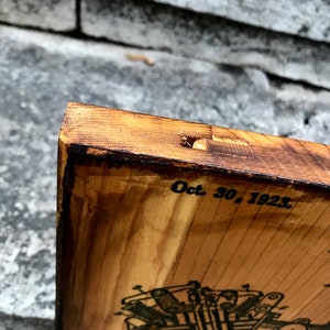 Puede incluir: Un letrero de madera con una ilustraci&oacute;n en blanco y negro de un motor de motocicleta. La fecha "Oct. 30, 1923" est&aacute; impresa en el letrero.