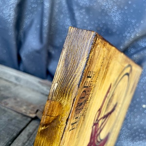 May include: A wooden sign with a vintage design featuring the text "Heindee" and a stylized illustration of a bicycle. The sign has a dark brown stain and a distressed look.