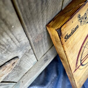 May include: A wooden sign with a vintage illustration of an Indian motorcycle. The sign is painted in shades of brown and red. The text on the sign reads "Indian" and "World's Most Complete Motorcycle Line".