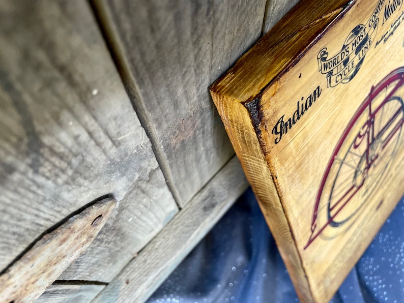 May include: A wooden sign with a vintage illustration of an Indian motorcycle. The sign is painted in shades of brown and red. The text on the sign reads "Indian" and "World's Most Complete Motorcycle Line".