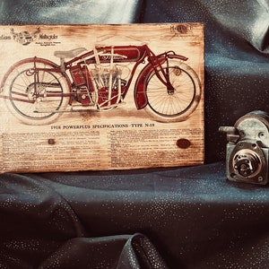 May include: A vintage wooden sign with a black and white illustration of a red 1918 Indian Motorcycle. The sign has the text "Indian Motorcycles" and "1918 Powerplus Specifications - Type N-18" printed on it.