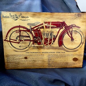 May include: A vintage wood sign with a detailed illustration of a red 1918 Indian Powerplus motorcycle. The sign includes the text "World's Most Copied Motorcycle" and "Indian Motorcycle Company Springfield, Mass."