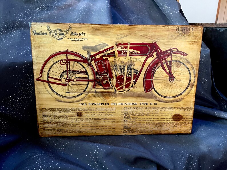 May include: A vintage wood sign with a detailed illustration of a red 1918 Indian Powerplus motorcycle. The sign includes the text "World's Most Copied Motorcycle" and "Indian Motorcycle Company Springfield, Mass."