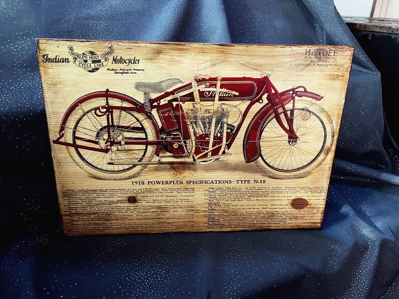 May include: A vintage advertisement for an Indian motorcycle, featuring a red motorcycle with a black engine and white wheels. The text on the advertisement reads "Indian World's Most Copied Cycle Line Motorcycles" and "1918 Powerplus Specifications - Type N-18".