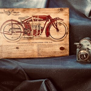 May include: A vintage wood plaque with a red and black illustration of a 1918 Indian Motorcycle, model N-18. The plaque features the text "Indian Motorcycles" and "1918 Powerplus Specifications - Type N-18".