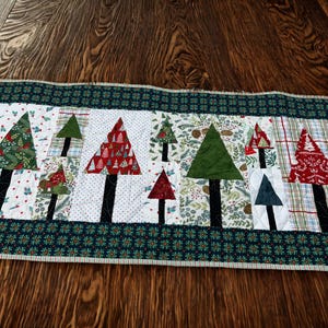 May include: A rectangular Christmas-themed quilted table runner. The runner features a white center with appliqued Christmas trees in various colors and patterns. The border is a dark green floral print with a striped edge.