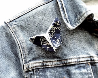 May include: A navy blue and white patterned butterfly brooch pinned to a denim jacket. The butterfly is made of fabric and has a delicate, origami-like appearance. The denim jacket is a light blue wash, and the brooch is the focal point.