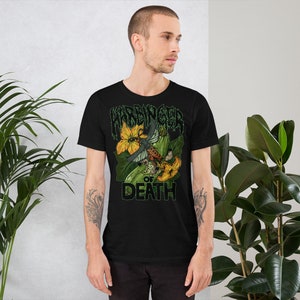 May include: Black t-shirt featuring a graphic design with the text "HARBINGER OF DEATH". The design includes a detailed illustration of a moth with orange and brown markings, set against yellow flowers and green foliage. The shirt is made of a soft material.