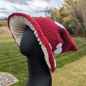 Puede incluir: Un sombrero de ganchillo con forma de seta, con una copa de color rojo intenso y una parte inferior ancha de color crema. El exterior de la copa roja presenta varios parches circulares de ganchillo blanco. El sombrero se exhibe sobre un maniquí negro.