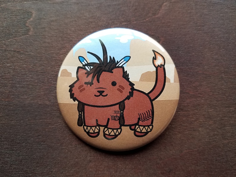 May include: A round button featuring a cartoon illustration of a brown cat wearing a Native American headdress. The cat is wearing moccasins and has a feather headdress. The background is a desert scene with a blue sky.