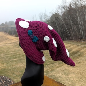 May include: A handmade, burgundy crochet hat with large white polka dots and a small teal and purple bow. The hat is displayed on a black mannequin head, showcasing its unique design and craftsmanship. The hat is a whimsical accessory.