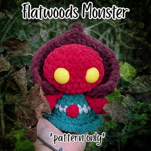 May include: A crocheted Flatwoods Monster plush toy featuring a round red head, large yellow eyes, and a dark maroon hood. The body is teal with a white zigzag trim and a small red heart detail. The text reads Flatwoods Monster and pattern only.