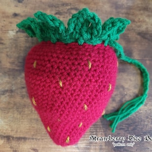 May include: A red crocheted strawberry-shaped pouch with green crocheted leaves at the top and yellow embroidered seeds. A green crocheted drawstring cord hangs from the side. The text "Strawberry Dice Bag pattern only" is visible.