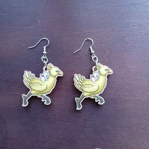 Op de afbeelding: Een paar oorbellen met een gele Chocobo-vogel met een wit wezen op zijn rug. De oorbellen hebben zilverkleurige oorhaken. De Chocobo heeft blauwe ogen en rent. De oorbellen zijn gemaakt van hout.