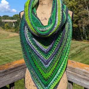 May include: A handmade, triangular crochet scarf in shades of green, gray, and black. The scarf is displayed on a mannequin, showcasing its intricate stitch pattern and color gradient. The background features a sunny outdoor scene.