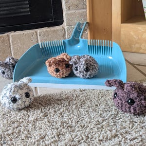 May include: Five handmade crochet bunny toys in various colours: brown, grey, white, and purple. The bunnies have black eyes and are displayed on a blue dustpan and a beige carpet.