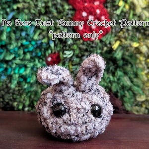 May include: A grey and white crocheted bunny with black eyes. The bunny is sitting on a brown surface. The text "No Sew Dust Bunny Crochet Pattern *pattern only*" is above the bunny.