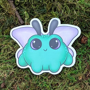 May include: A whimsical sticker featuring a cartoonish teal creature with large, purple-rimmed eyes and small antennae. The creature has lavender wings with glitter accents. The sticker is set against a backdrop of green moss.