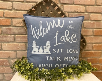May include: A square blue throw pillow featuring white printed text that reads Welcome to the Lake, SIT LONG, TALK MUCH, LAUGH OFTEN, accompanied by a graphic illustration of two Adirondack chairs positioned on either side of a small fire pit.