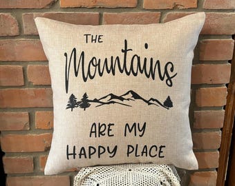 May include: A beige throw pillow with black text that reads "The Mountains Are My Happy Place". The pillow features a black silhouette of mountains and trees.