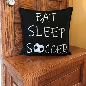 K&ouml;nnte beinhalten: Schwarzes Kissen mit wei&szlig;em Text "Eat Sleep Soccer" und einer wei&szlig;en Fu&szlig;ballgrafik.