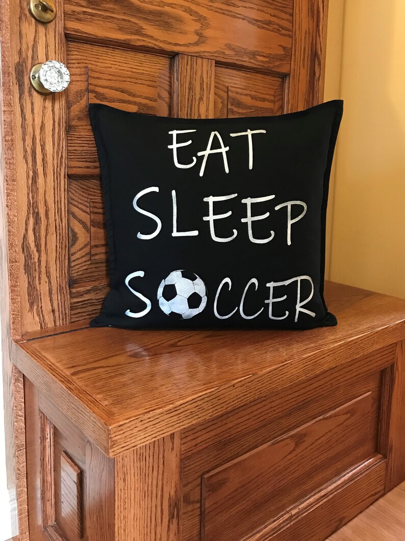 K&ouml;nnte beinhalten: Schwarzes Kissen mit wei&szlig;em Text "Eat Sleep Soccer" und einer wei&szlig;en Fu&szlig;ballgrafik.