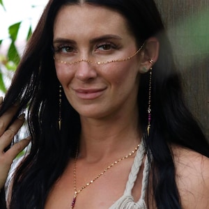 Può includere: Una donna indossa gioielli d'oro, tra cui una catena per il viso, orecchini, una catena per la mano e una collana con pendente. Indossa un abito beige con scollatura profonda. Lo sfondo presenta un rigoglioso fogliame verde e un tronco d'albero.