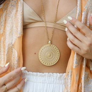 Puede incluir: Un collar largo dorado con un colgante grande y redondo con un diseño intrincado y detallado. El colgante cuelga de una cadena delicada. La persona lleva un sujetador dorado y un chal transparente naranja con detalles plateados.