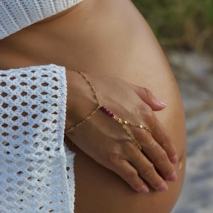 May include: A gold chain bracelet with a small red gemstone accent. The bracelet is worn on the wrist and has a finger ring attached to it.