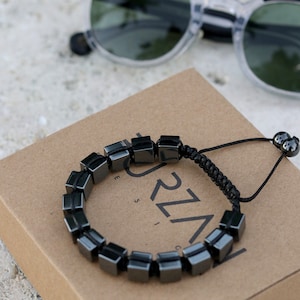 May include: A black hematite bracelet with square beads and a black cord. The bracelet is on a brown box with the word "REVEAL" printed on it.