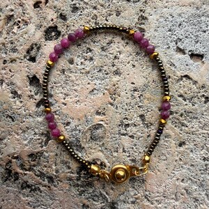 May include: A beaded bracelet featuring small, round, deep pink gemstones interspersed with gold-colored beads and dark metallic beads. The bracelet has a gold-colored clasp. The jewelry is displayed on a textured, neutral-toned surface.