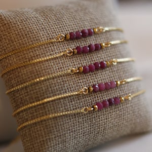 Puede incluir: Seis delicados brazaletes de cadena dorada con pequeñas cuentas de piedras preciosas de color rosa oscuro. Los brazaletes se muestran sobre una superficie de arpillera marrón.