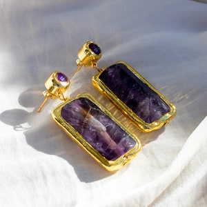May include: Gold-toned earrings featuring rectangular, deep purple amethyst stones. The earrings have a gold border and a small purple stone at the top. The earrings are displayed on a white, textured fabric.