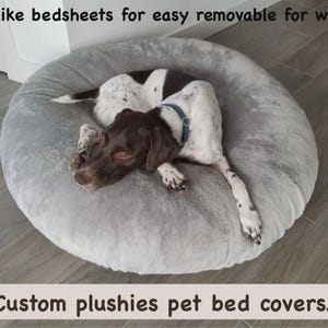 Puede incluir: Una funda redonda de felpa gris claro para cama de mascota con una superficie suave y texturizada. El texto "Made like bedsheets for easy removable for washing" y "Custom plushies pet bed covers" es visible. Un perro manchado descansa.