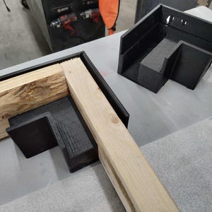 May include: A close-up of a wooden frame being assembled with black plastic corner brackets. The brackets are designed to create a 90-degree angle.