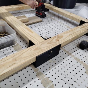 May include: A partially constructed wooden frame with black metal corner brackets. A red and black cordless drill rests on the frame. The frame is on a white pegboard surface.